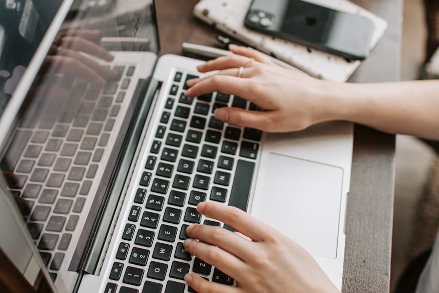 From above of unrecognizable woman sitting at table and typing on keyboard of computer during remote work in modern workspace